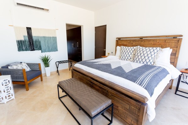 A bedroom with a wooden bed, a bench, a small table, a potted plant, and a wall-mounted tapestry.