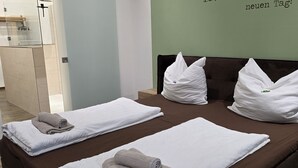 Habitación de hotel con dos camas, cada una con ropa blanca y toallas enrolladas. Se ve una pared verde con texto en alemán.