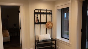 A corner room with a black metal rack holding a hat and a white pillow, a window, and a wooden floor.