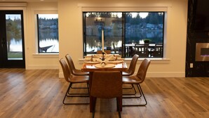 A dining area with a wooden table and chairs, large windows with a view of a lake and buildings, and a modern kitchen with a built-in oven.