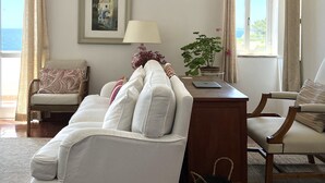 Een woonkamer met een witte bank, een houten bijzettafeltje met een lamp, een ingelijst schilderij aan de muur en uitzicht op de zee door een raam.