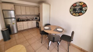 Eine kompakte Küche mit Kühlschrank, Mikrowelle und Herd. Ein Esstisch mit zwei Stühlen und eine Uhr hängen an der Wand.