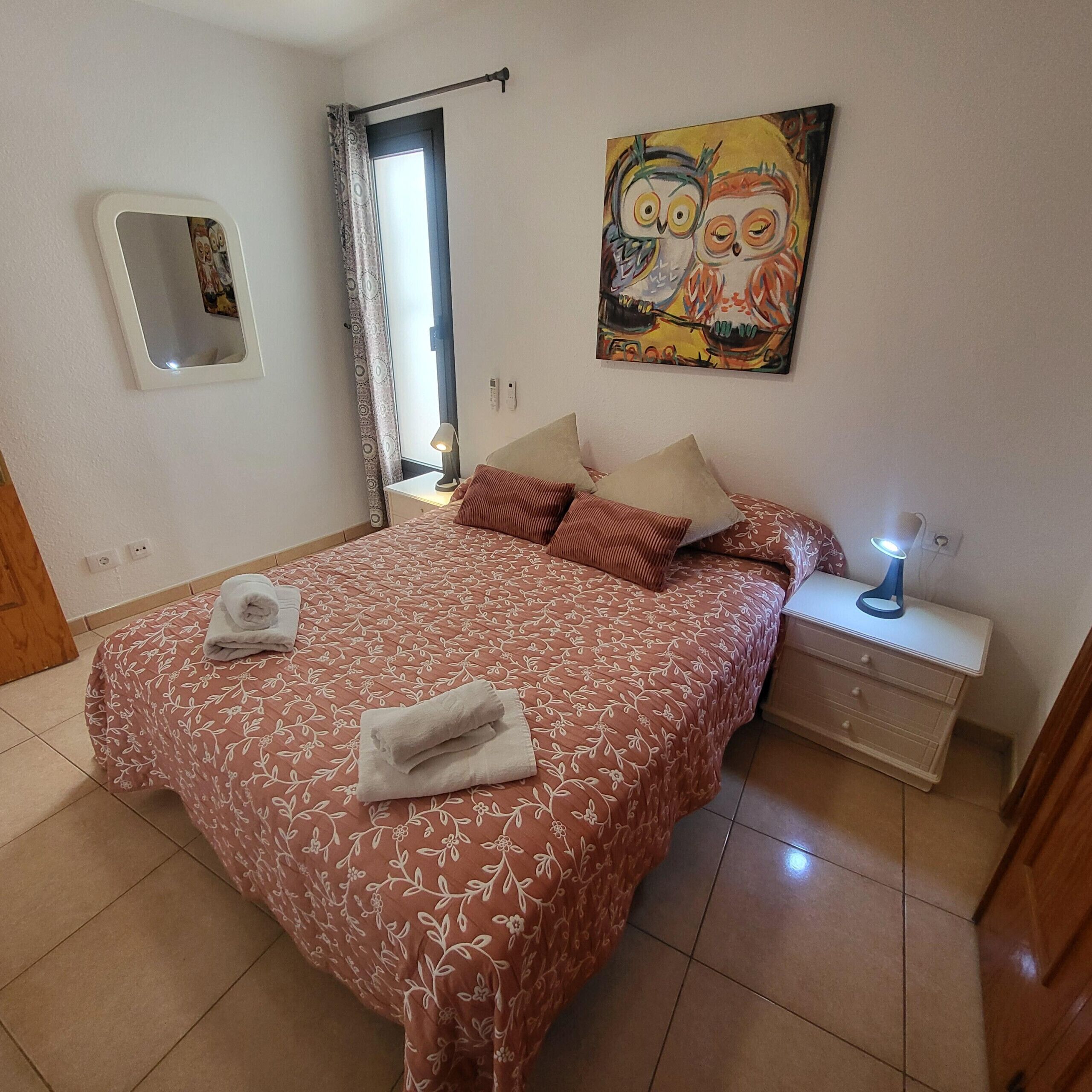 A bedroom with a bed, pillows, a nightstand, a lamp, a painting, a window with curtains, and a mirror.