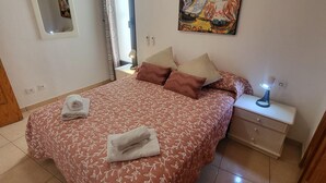 Ein Schlafzimmer mit Bett, Kissen, Nachttisch, Lampe, einem Gemälde, einem Fenster mit Vorhängen und einem Spiegel.