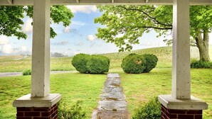 A covered walkway with brick columns, leading to a grassy area with two neatly trimmed bushes and a stone path.