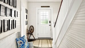 A hallway with a white door, framed pictures on the wall, a wooden chair, and a patterned rug.