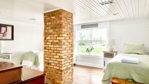 A room with a brick fireplace, a bed with a green bedspread, a nightstand with a lamp, and a window with a view of greenery.