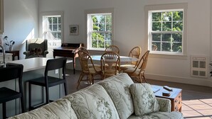 Un salon spacieux avec un grand canapé à motifs floraux, un coin repas avec des chaises en osier et de grandes fenêtres laissant entrer la lumière naturelle.