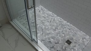 Une cabine de douche en verre incurvé avec un puits de lumière, des murs recouverts de carreaux blancs et un sol à motifs.