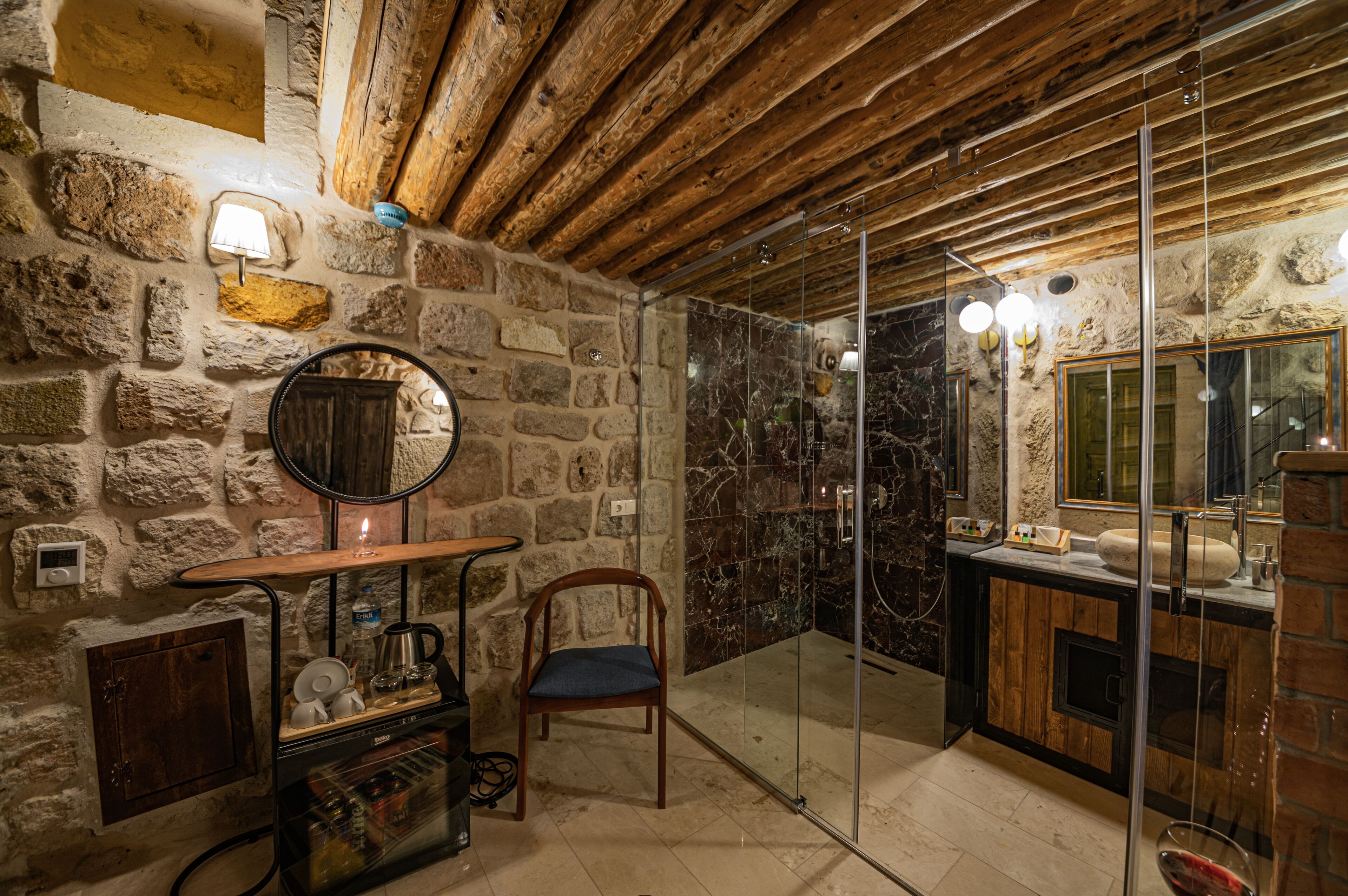 A rustic bathroom with stone walls, wooden beams, a glass shower enclosure, and a wooden chair.