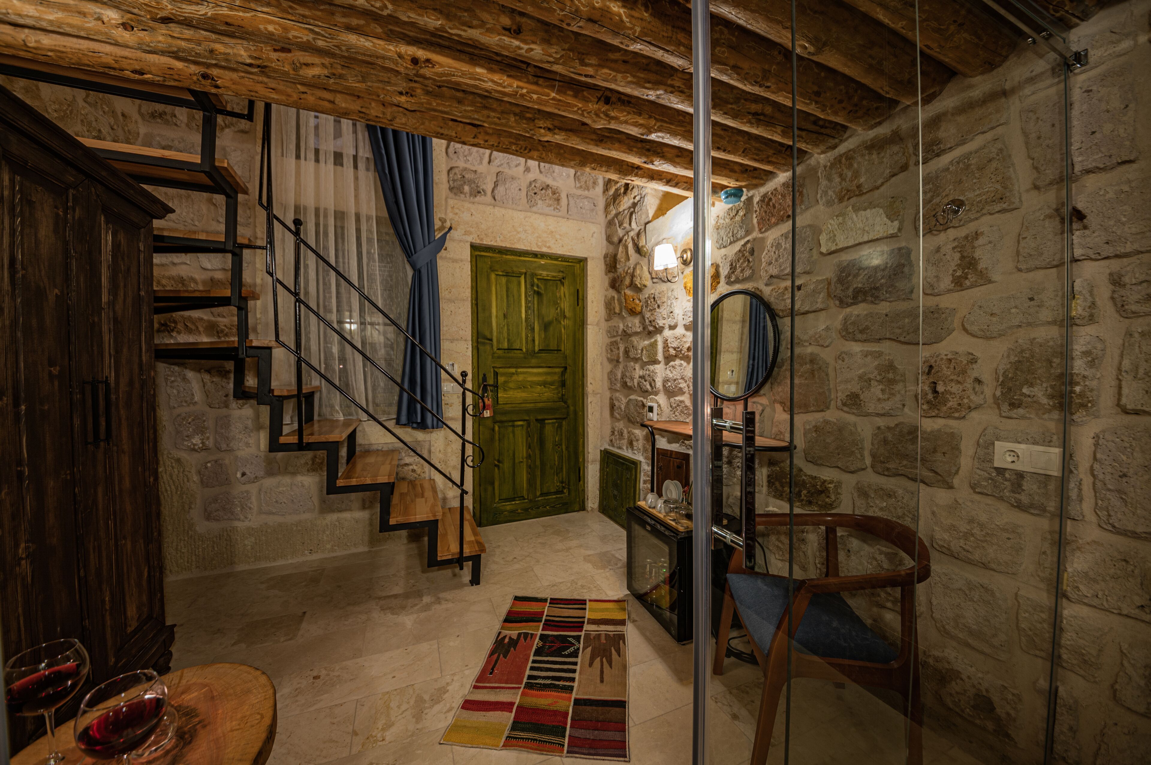 Un interior rústico con una escalera de madera, paredes de piedra, una puerta verde y una pequeña mesa con copas de vino.