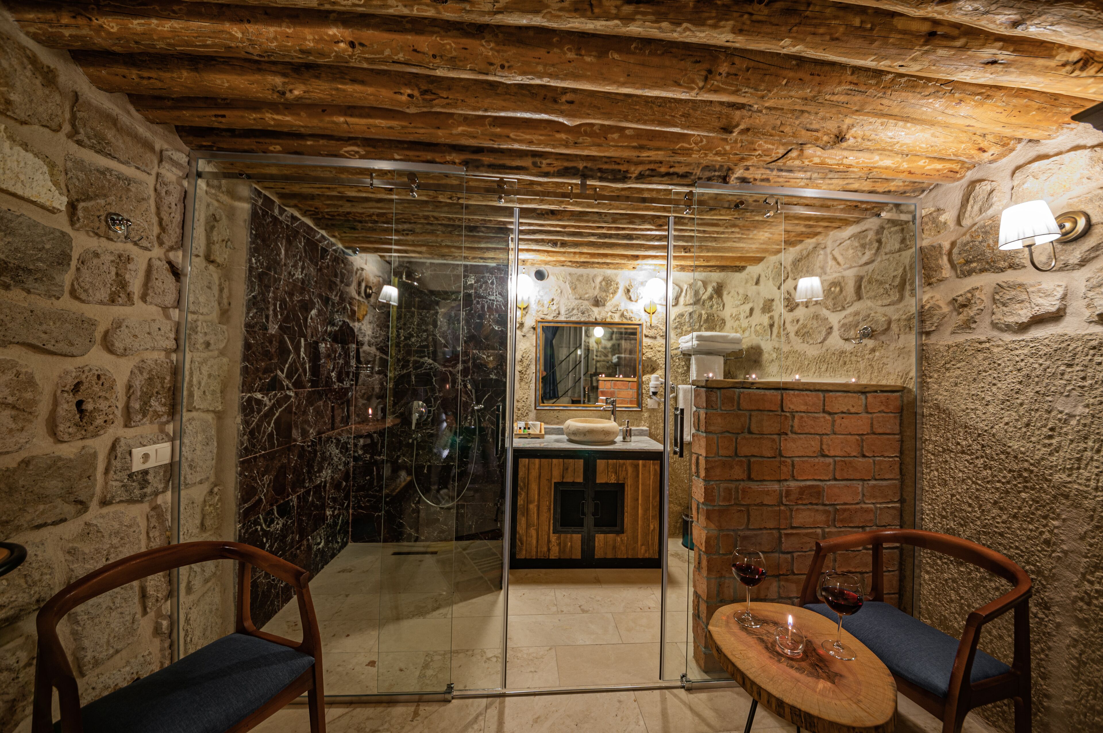 Un baño rústico con paredes de piedra, techo de madera, cabina de ducha con vidrio y tocador de madera con lavamanos y espejo.