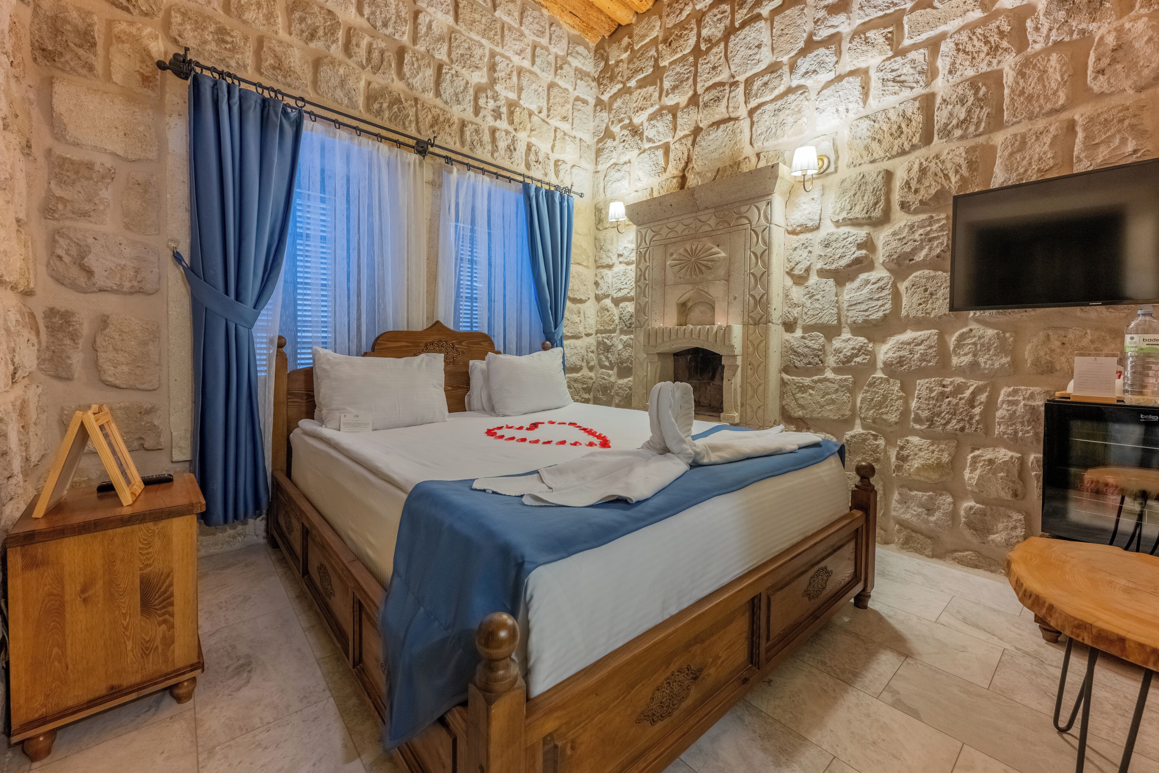 A stone-walled bedroom with a bed, curtains, a fireplace, a television, and a small wooden table.