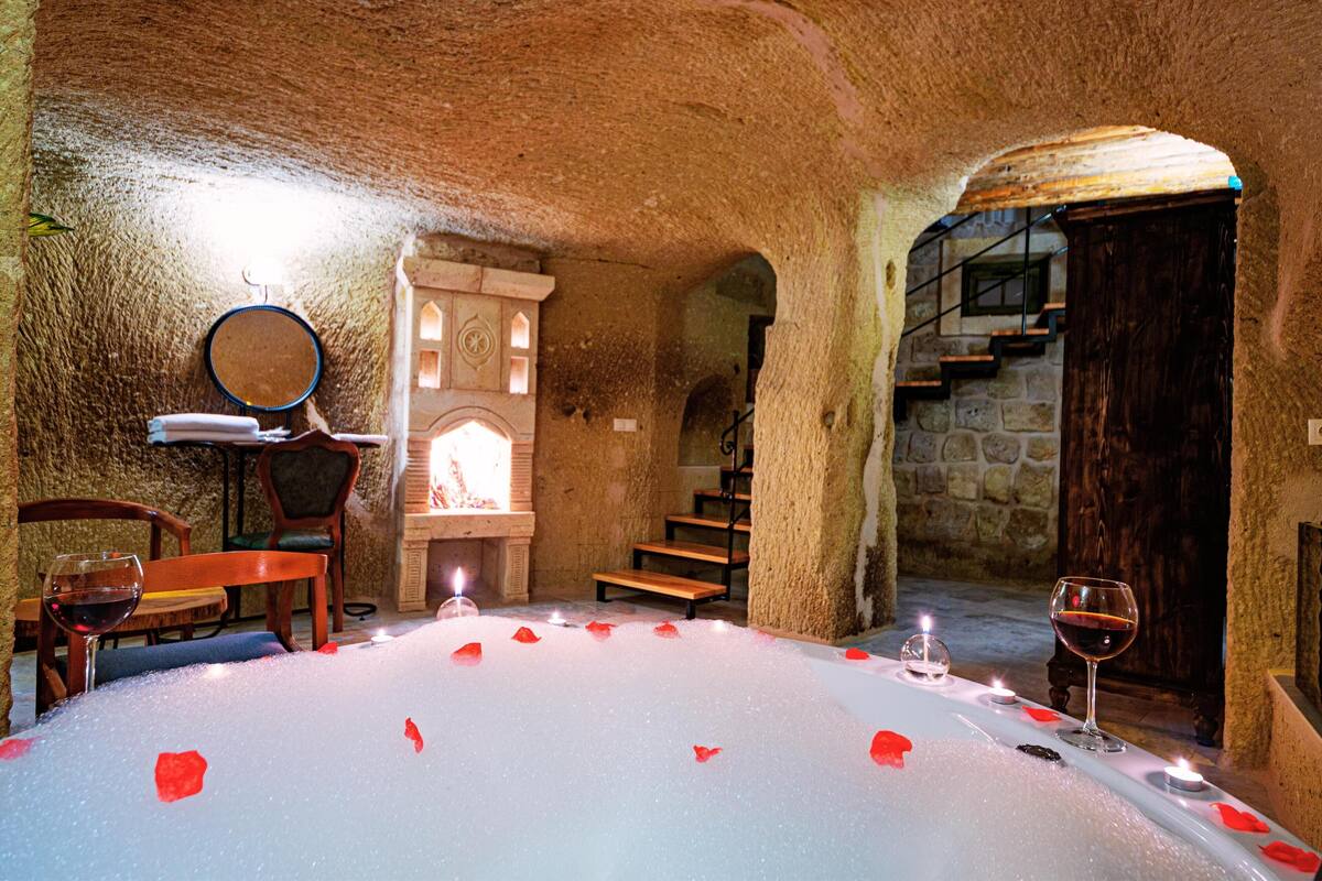 A cozy, candle-lit room with a stone interior, a wooden staircase, and a table set with rose petals and wine glasses.