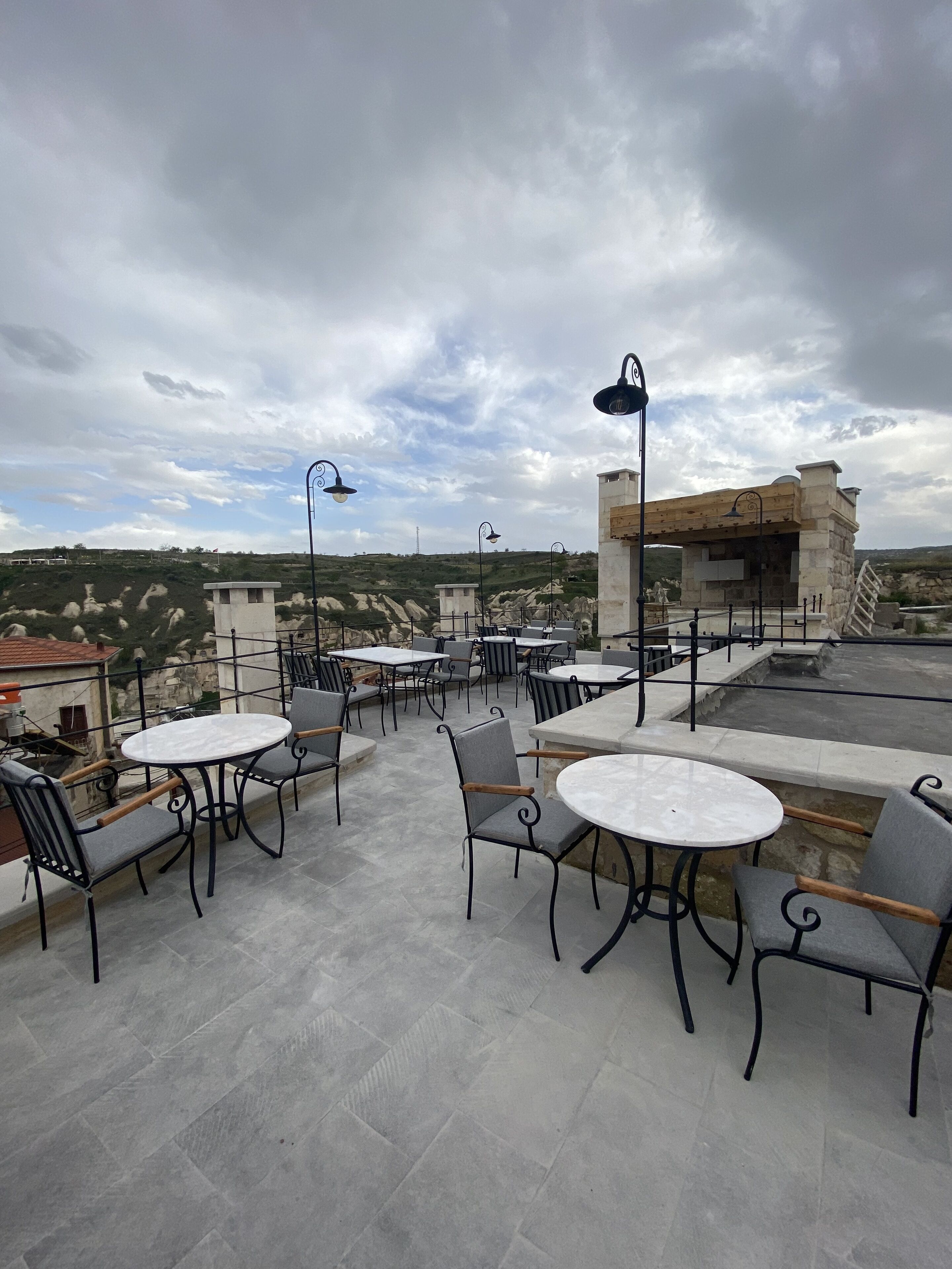 Um terraço ao ar livre com mesas e cadeiras redondas, paredes de pedra e vista para a paisagem circundante.