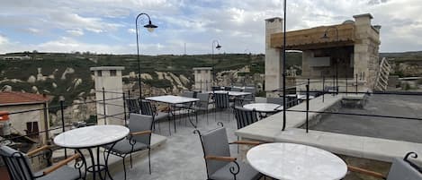 Um terraço ao ar livre com mesas e cadeiras redondas, paredes de pedra e vista para a paisagem circundante.