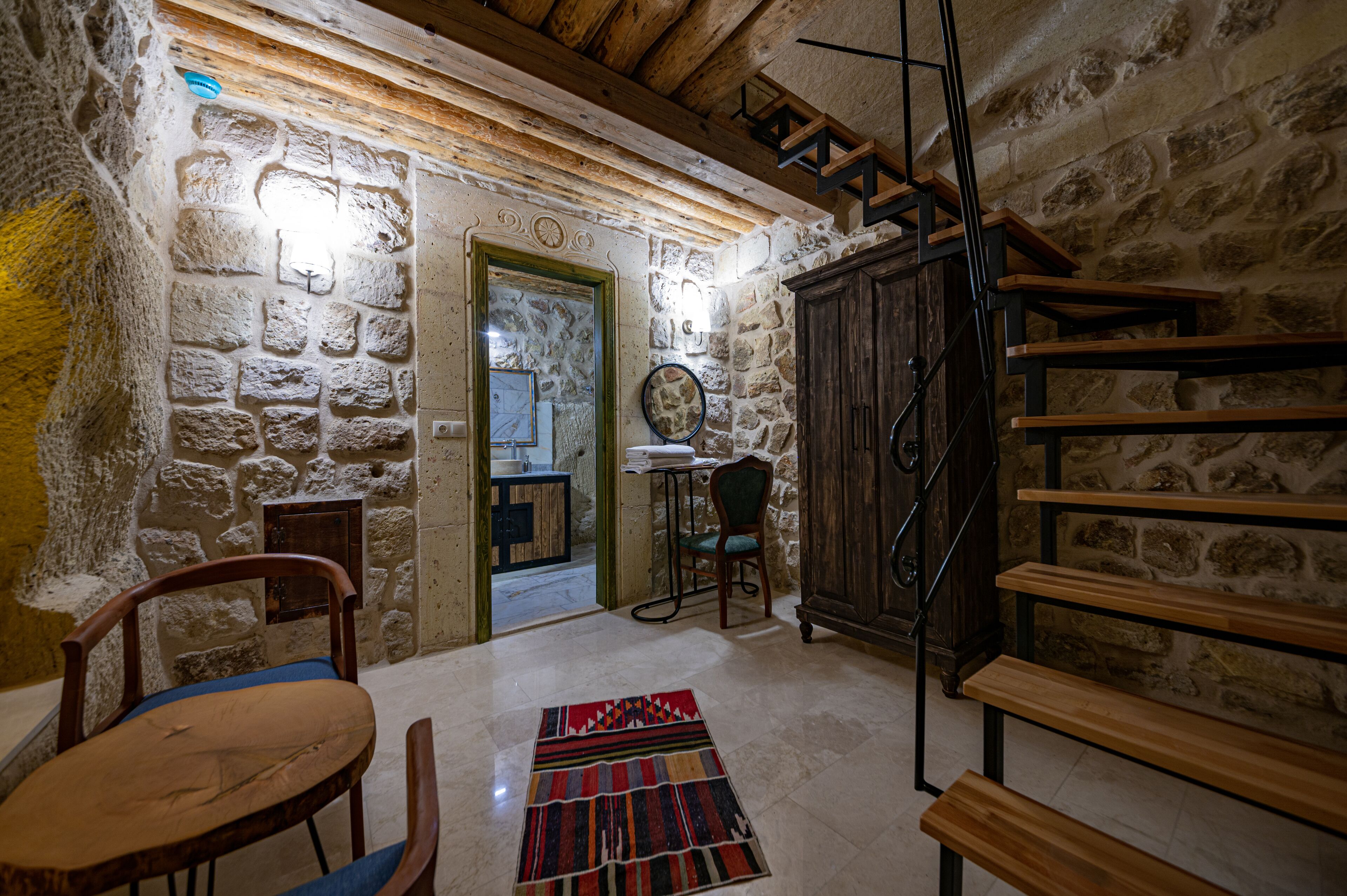 Una habitación rústica con paredes de piedra, un banco de madera, una silla, una mesita y una escalera.