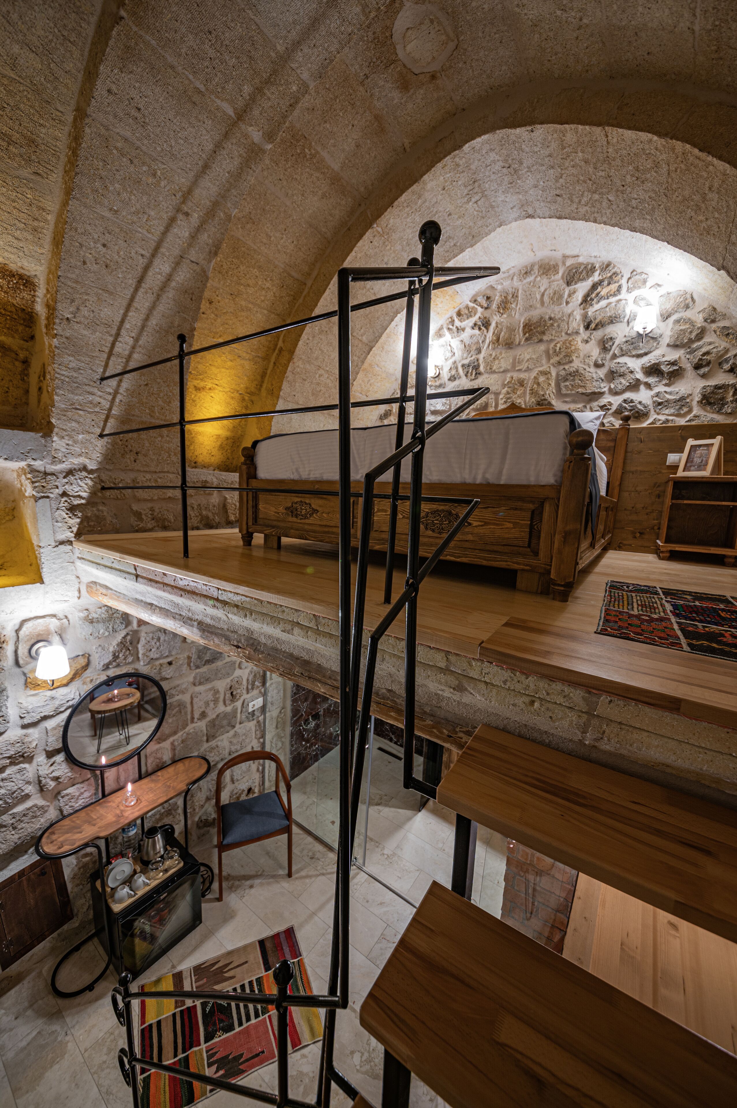 A rustic stone-walled room with a loft bed, a wooden table, a chair, and a small table with a lamp.