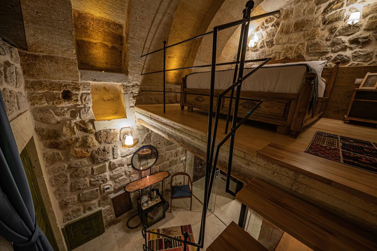 A rustic stone-walled room with a wooden bed, a small table with a lamp, and a chair.
