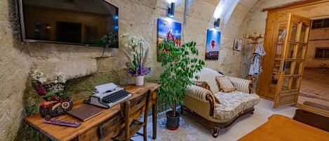 A cozy, rustic room with a wooden desk, a comfortable sofa, a television mounted on a stone wall, and a potted plant.
