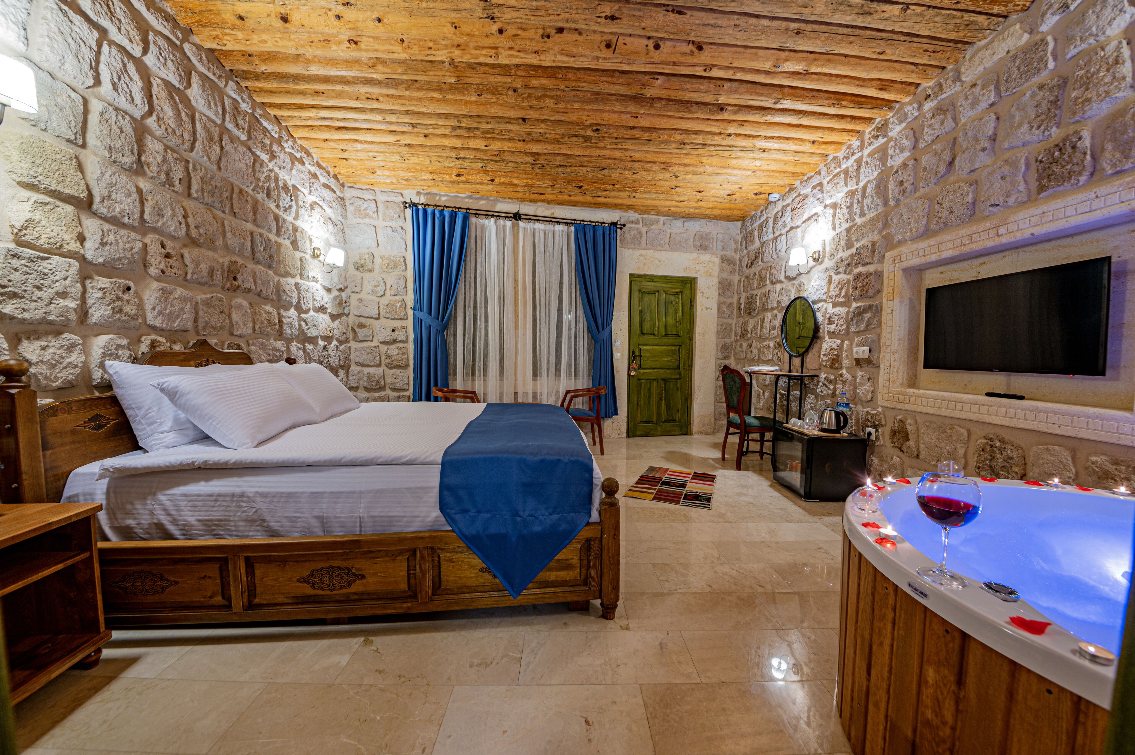 A stone-walled bedroom with a wooden bed, a stone fireplace, and a TV mounted on the wall.
