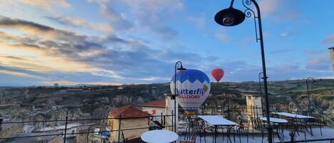Una terraza al aire libre con mesas y sillas, un anuncio de paseo en globo aerostático y vistas de un valle.