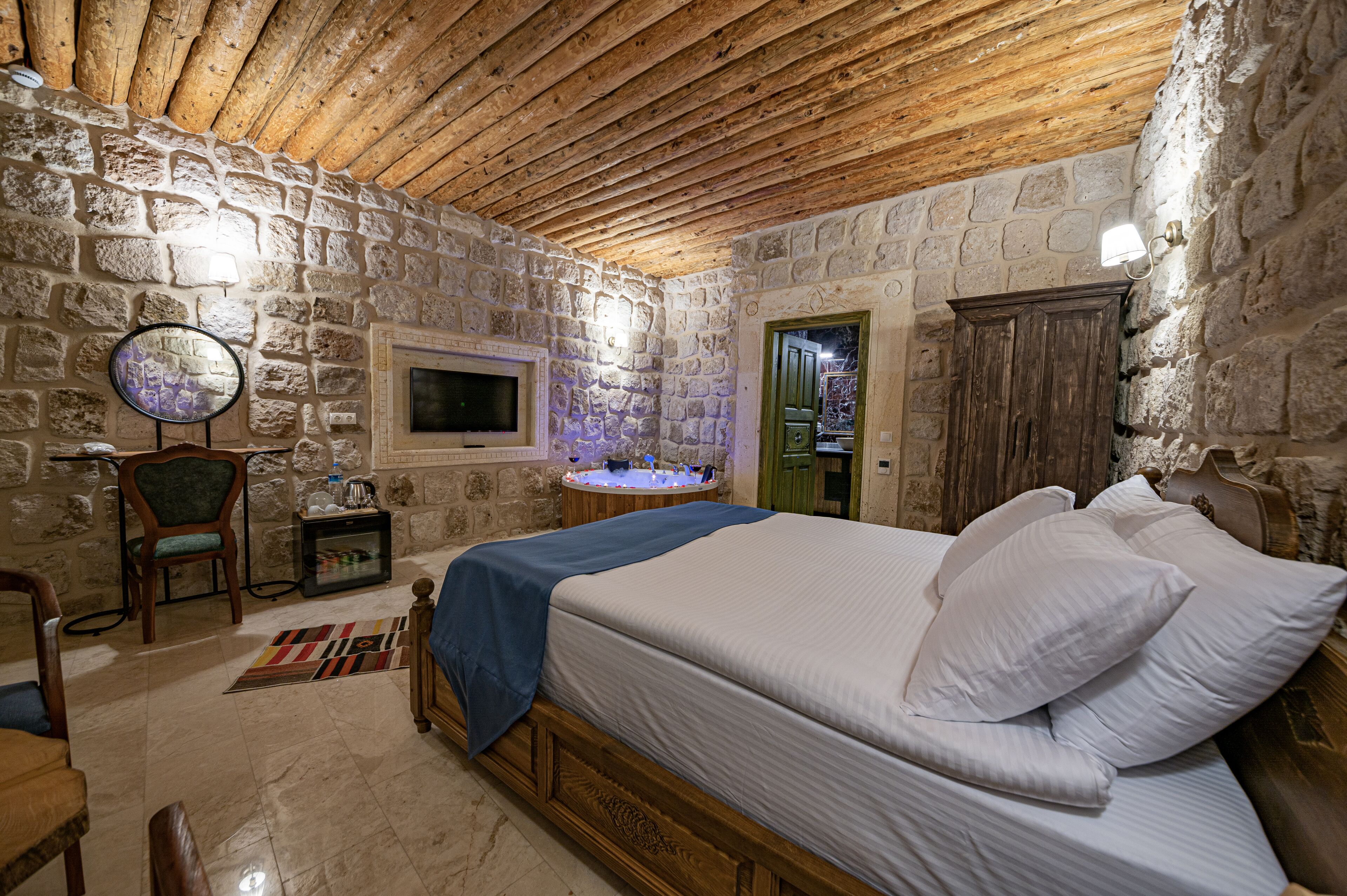 Une chambre aux murs de pierre et au plafond en bois, avec un lit recouvert de draps blancs, une petite table avec une lampe et un téléviseur fixé au mur.