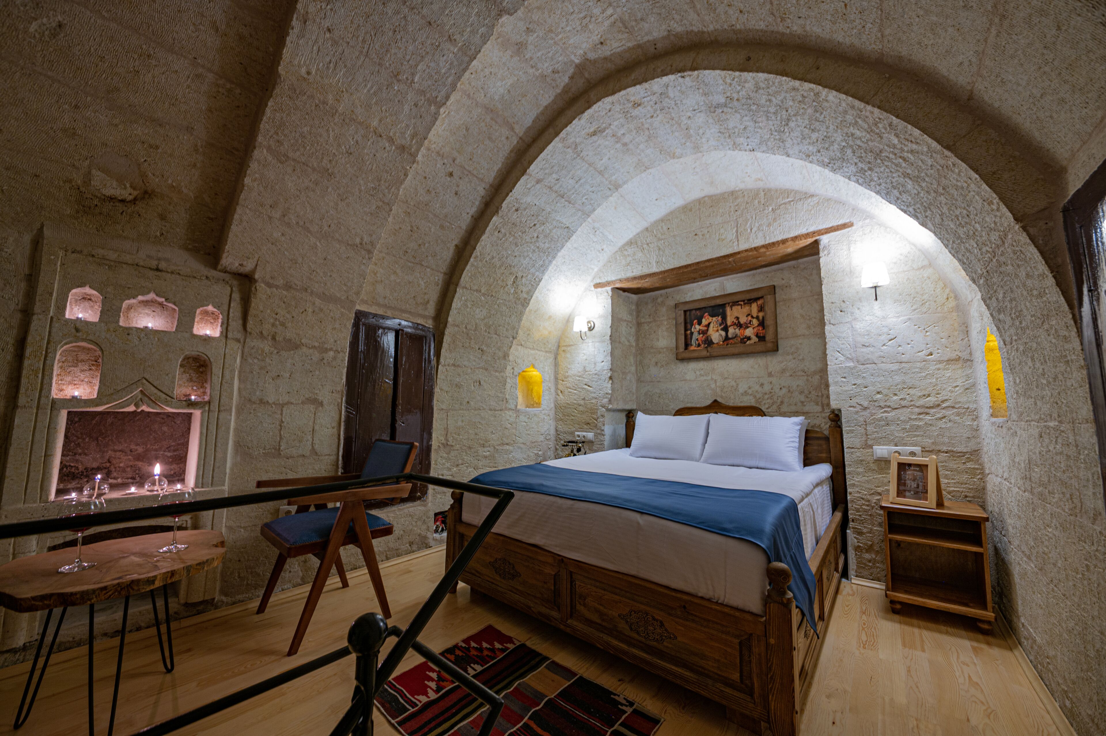A room with a stone arch ceiling, a bed with a wooden headboard, a small table with candles, and a chair.