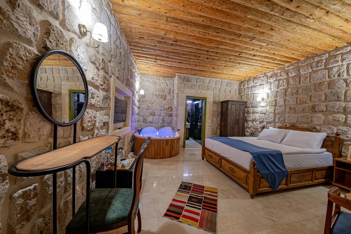 A stone-walled room with a wooden ceiling, a bed, a chair, a desk, and a bathtub with lights.