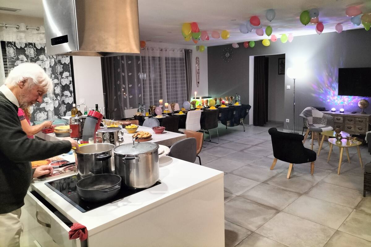 A kitchen with a stove, pots, and a person cooking. There is a dining area with a table set for a meal, balloons, and a globe on a stand.