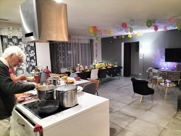 Una cocina con estufa, ollas y una persona cocinando. Hay un comedor con una mesa dispuesta para una comida, globos y un globo terráqueo sobre un soporte.