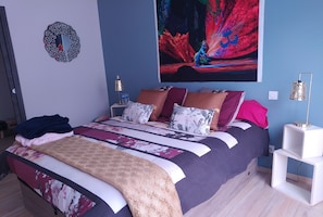 A bedroom with a bed, pillows, a blanket, a wall art piece, a side table, and a lamp.