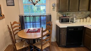 A dining area with a round table and wooden chairs, a chandelier, a window with curtains, and a kitchen with cabinets and appliances. - Sunrise Side Retreat - Sleeps 7 (Harrisville)