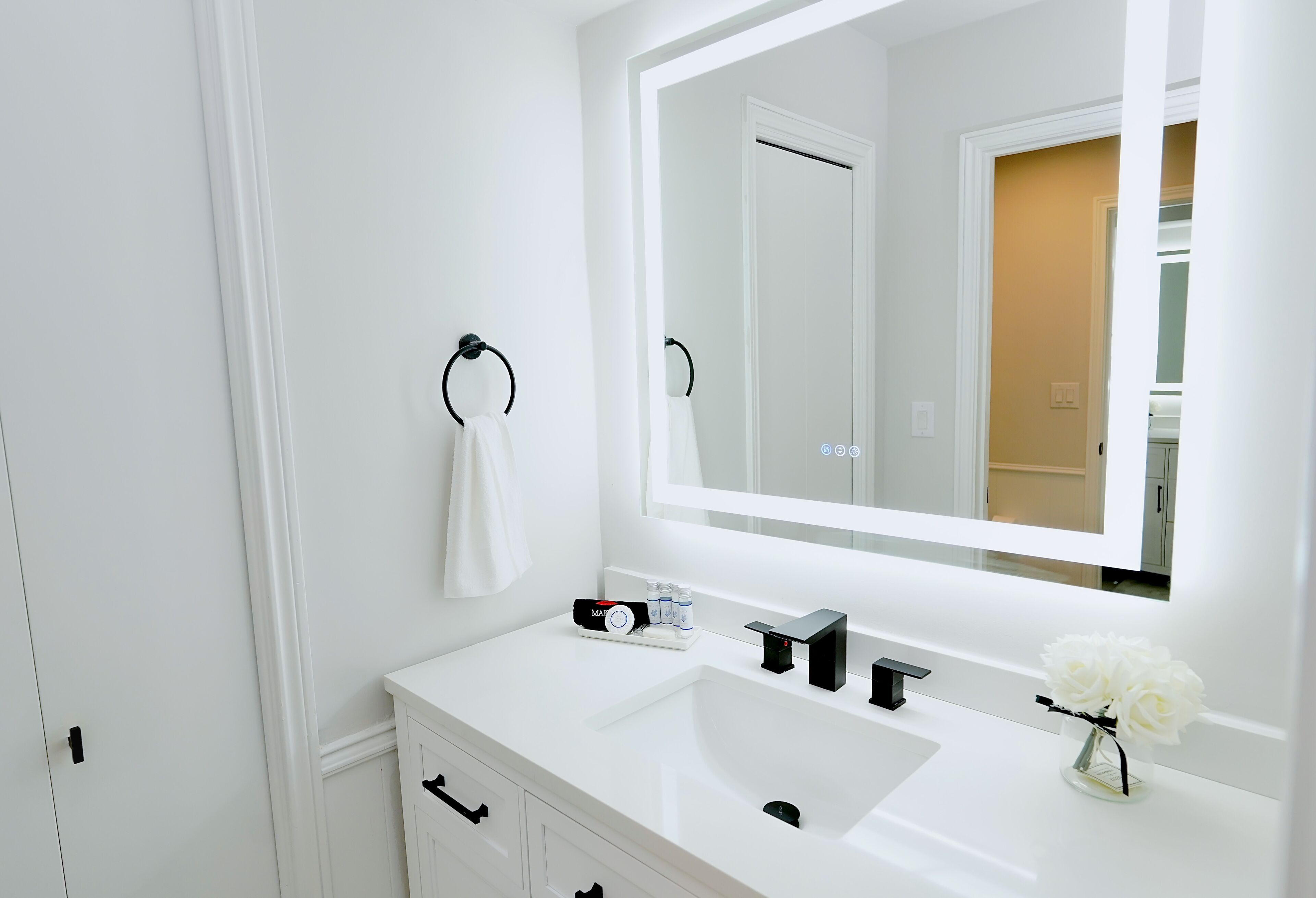 A modern bathroom with a white vanity, a large mirror, and a vase of white flowers.