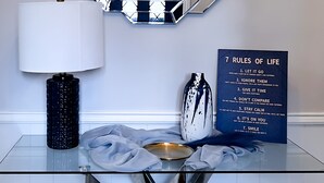 Ein modernes Interieur mit einem Glastisch, einer dekorativen Lampe, einem gerahmten Schild mit Lebensregeln und einem geometrischen Spiegel.