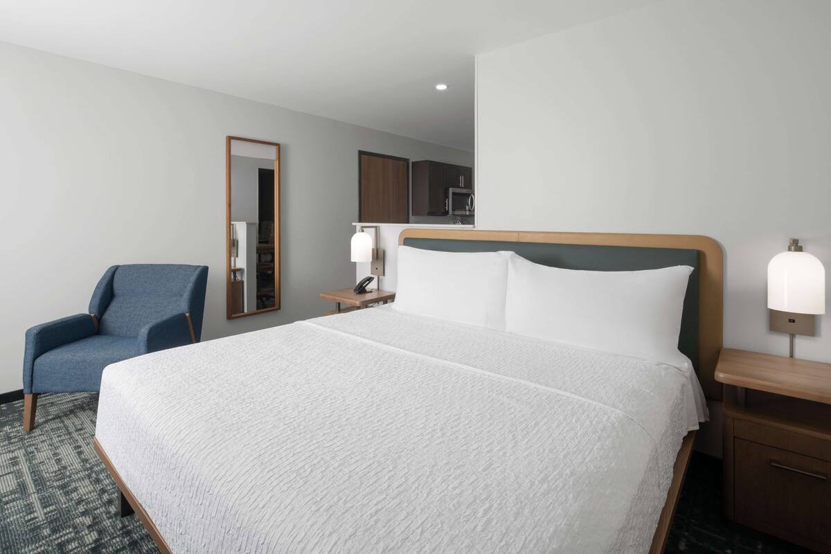 Ein Hotelzimmer mit einem großen Bett, einem Sessel, einem Nachttisch, einem Spiegel und einer kleinen Küchenzeile im Hintergrund.