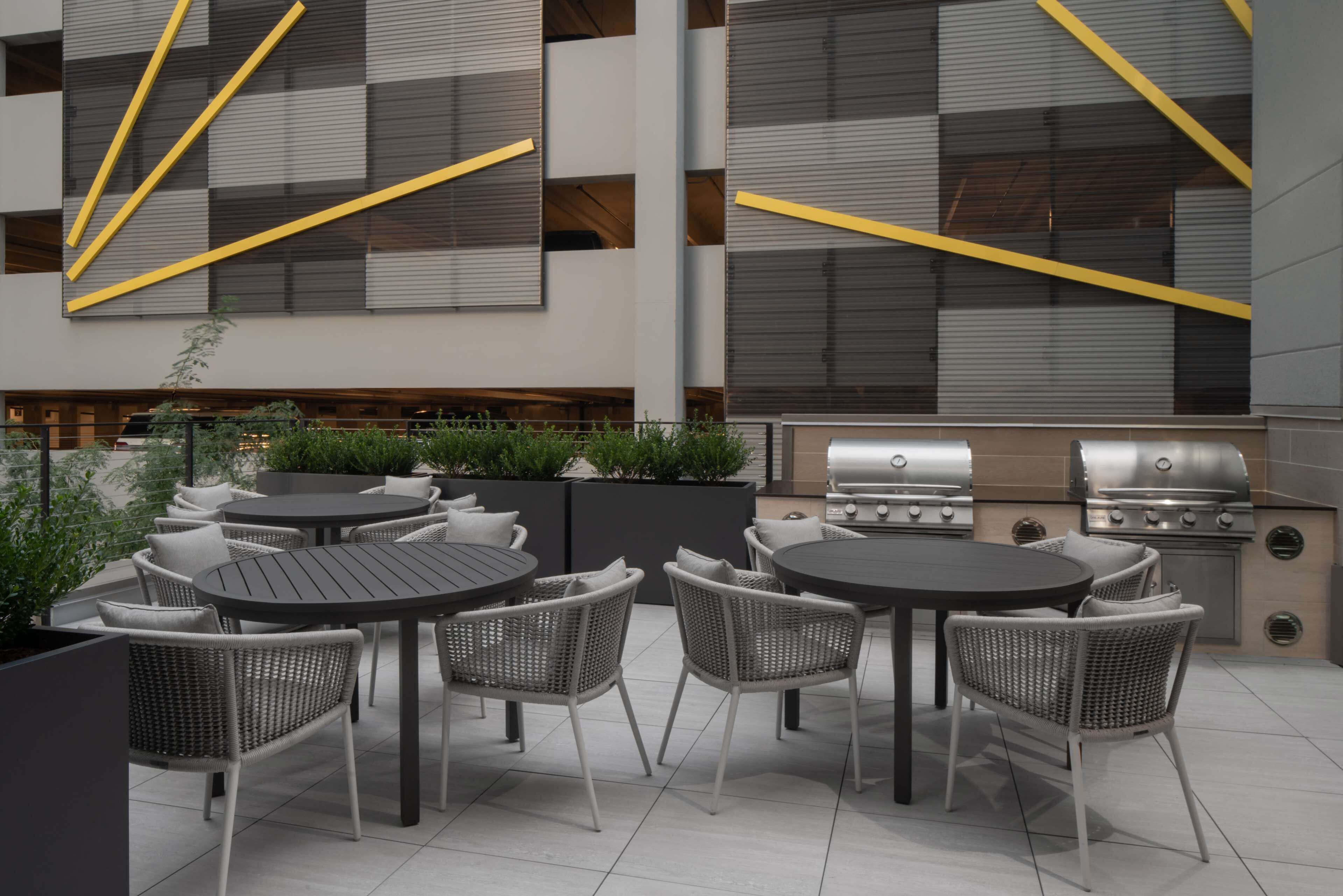 An outdoor patio with round tables and wicker chairs, a grill, and a modern building with geometric patterns.
