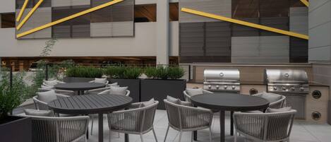 An outdoor patio with round tables and wicker chairs, a grill, and a modern building with geometric patterns.
