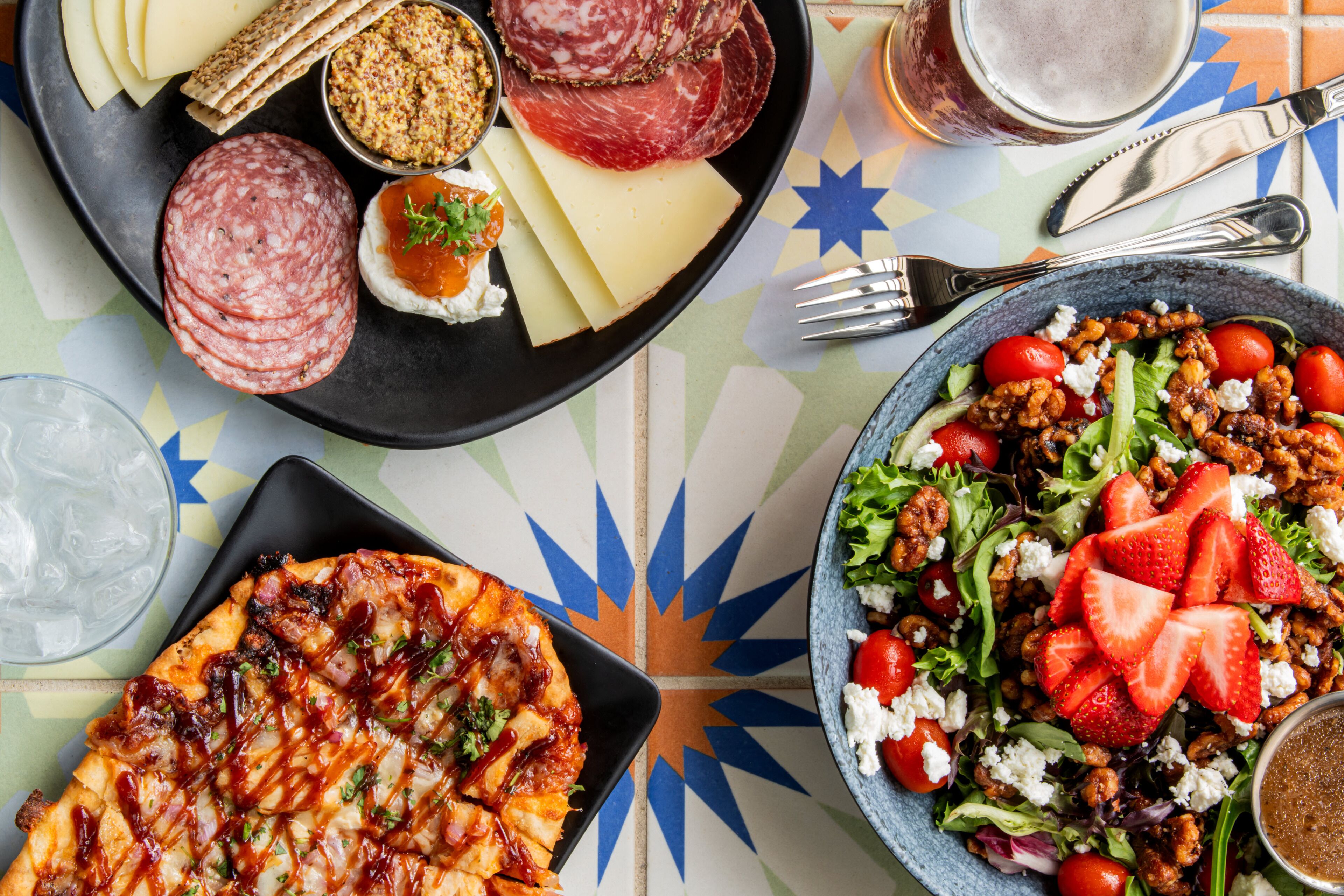 Variedad de alimentos, incluyendo pizza, ensaladas y quesos, sobre una mesa con fondo de azulejos.