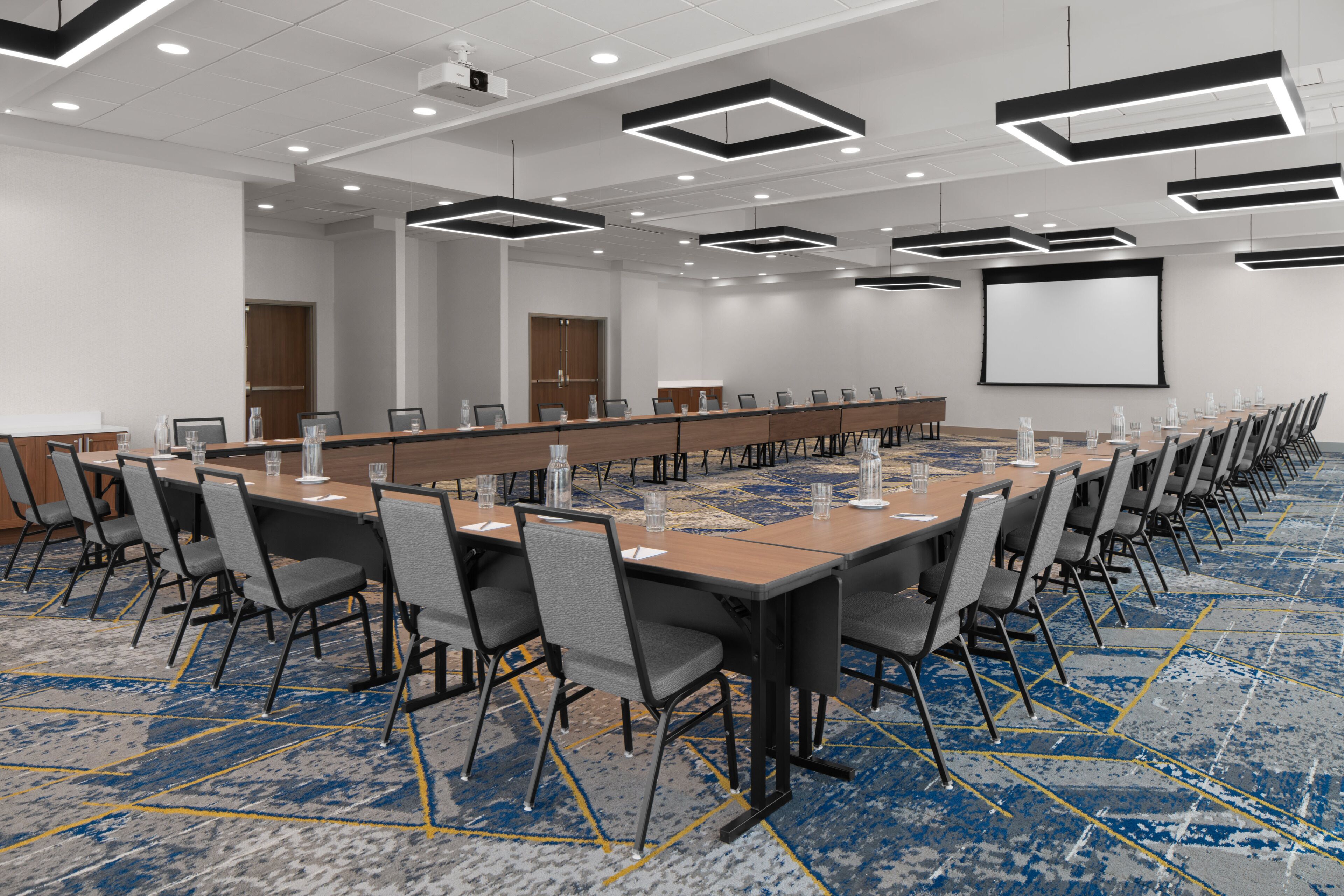 A conference room with a long table, multiple chairs, a projector screen, and a carpeted floor.