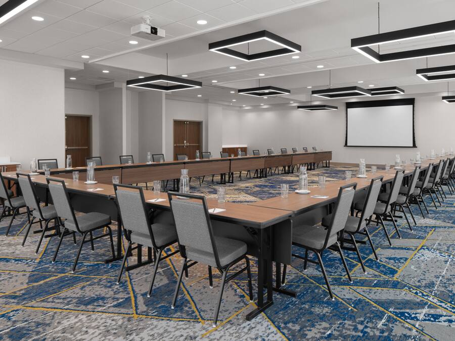 Un salón de conferencias con una larga mesa, varias sillas, una pantalla de proyector y piso alfombrado.