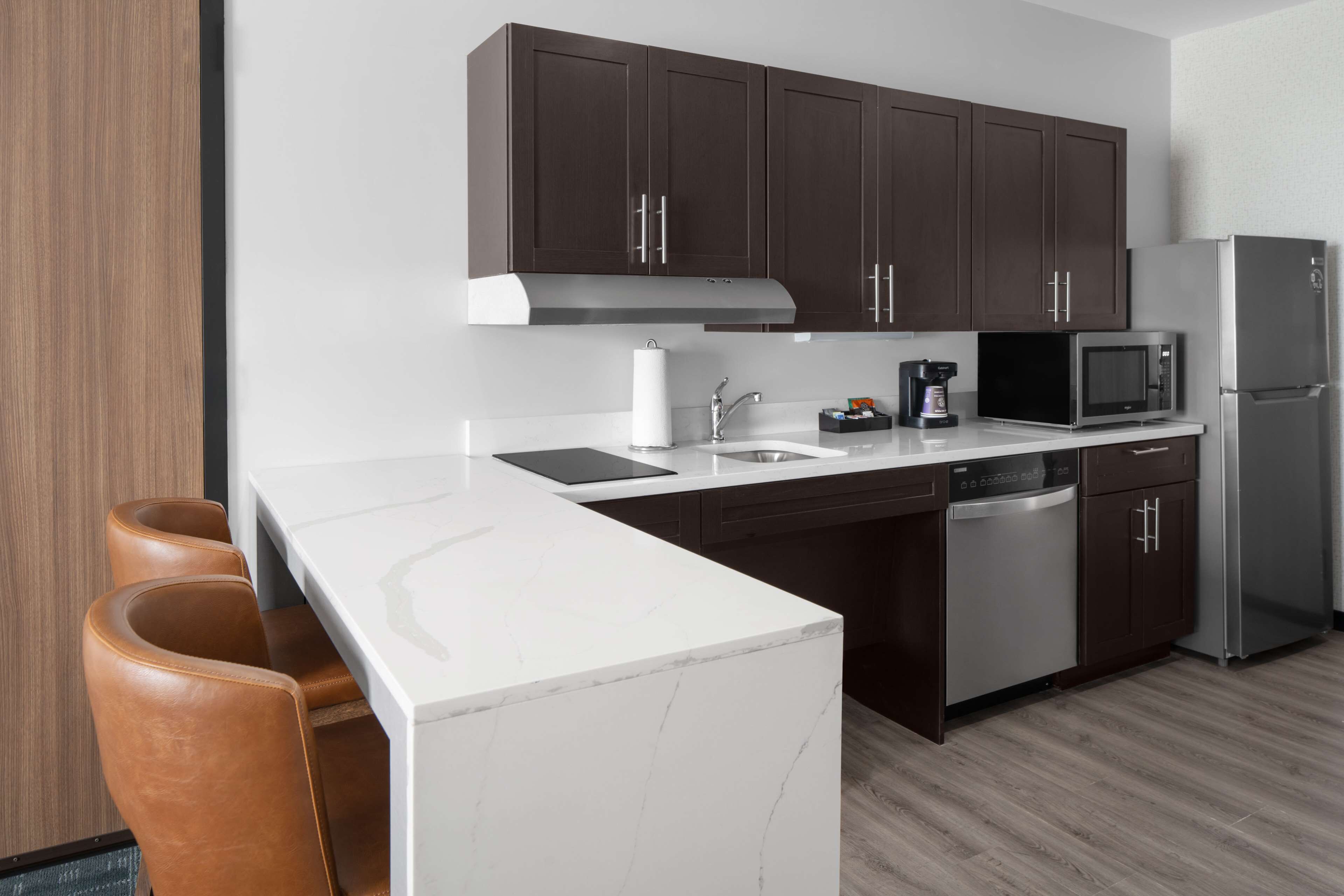 Una cocina moderna con armarios marrón oscuro, encimera blanca y electrodomésticos de acero inoxidable, incluyendo un microondas y un refrigerador.