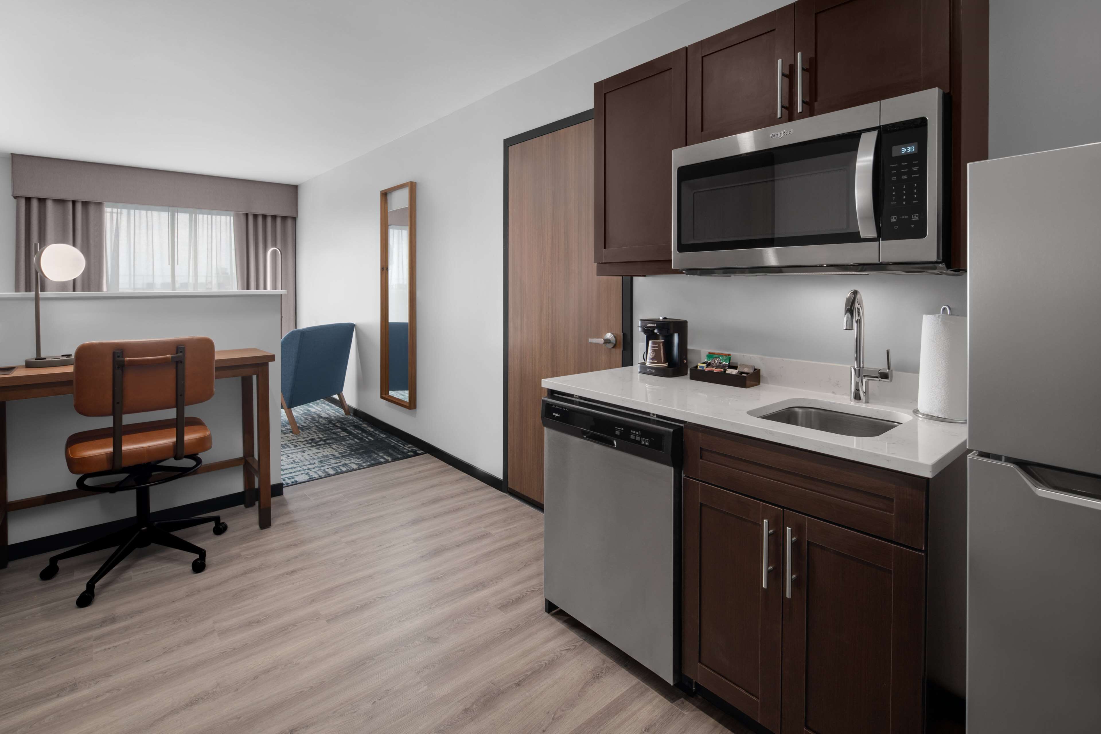 Habitación de hotel compacta con kitchenette, un escritorio y silla, un espejo grande y una ventana con cortinas.
