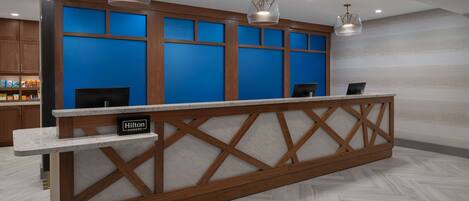 A hotel reception area with a wooden reception desk, blue backlit panels, and pendant lighting.