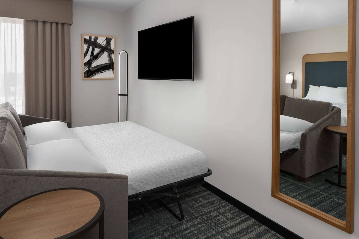 Ein Hotelzimmer mit Bett, Sessel, kleinem Tisch, Flachbildfernseher und Spiegel.