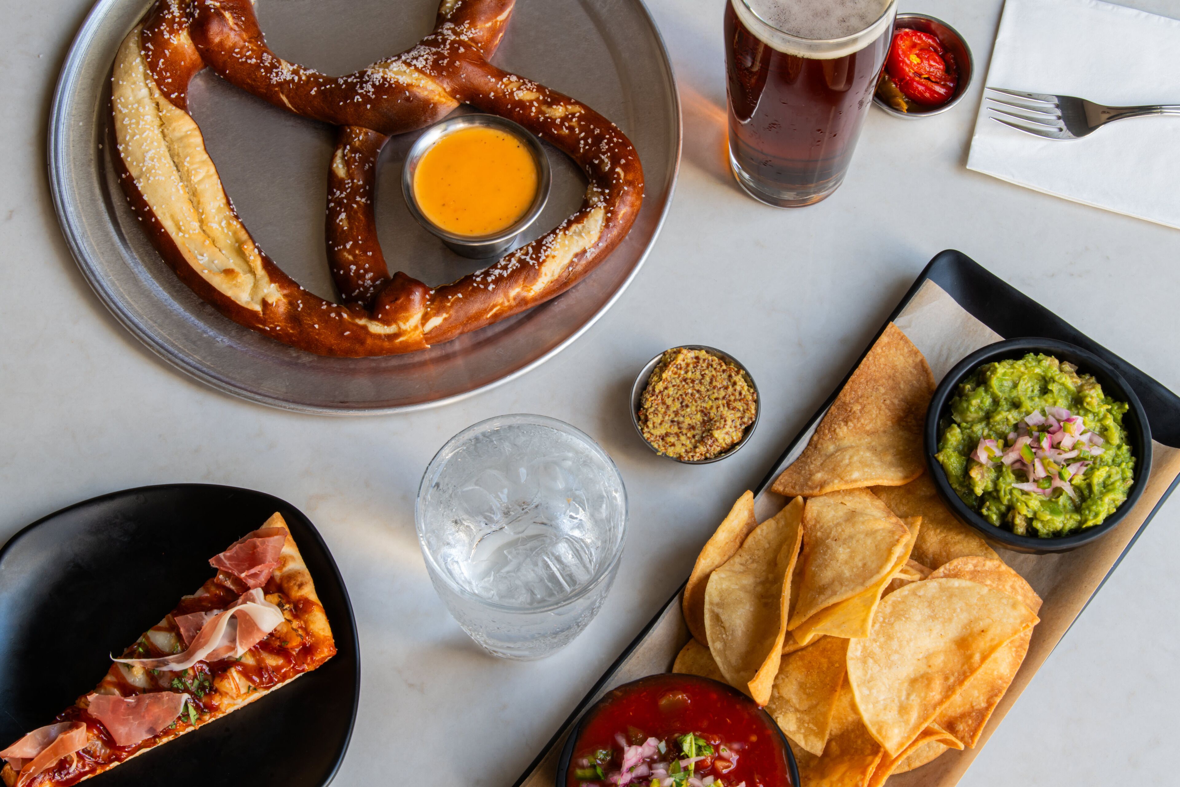 A table with a pretzel, a slice of pizza, a glass of beer, chips, and a bowl of guacamole.