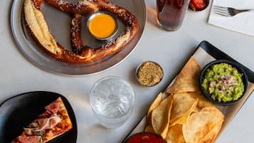 A table with a pretzel, a slice of pizza, a glass of beer, chips, and a bowl of guacamole.