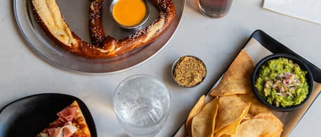 Una mesa con un pretzel, una porción de pizza, una jarra de cerveza, papas fritas y un tazón de guacamole.