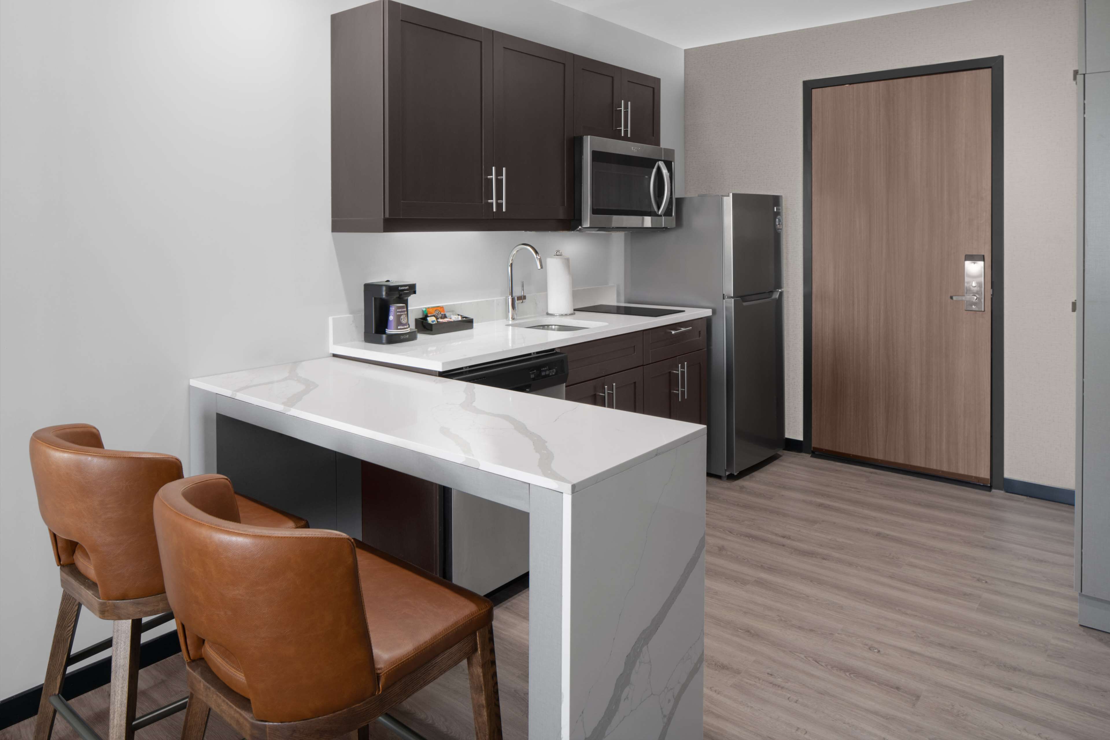 A modern kitchen with dark cabinets, a white countertop, a stainless steel refrigerator, and a microwave. There are two brown leather bar stools and a white sink.