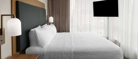 A hotel room with a large bed, a bedside table, a flat-screen TV mounted on the wall, and a window with curtains.